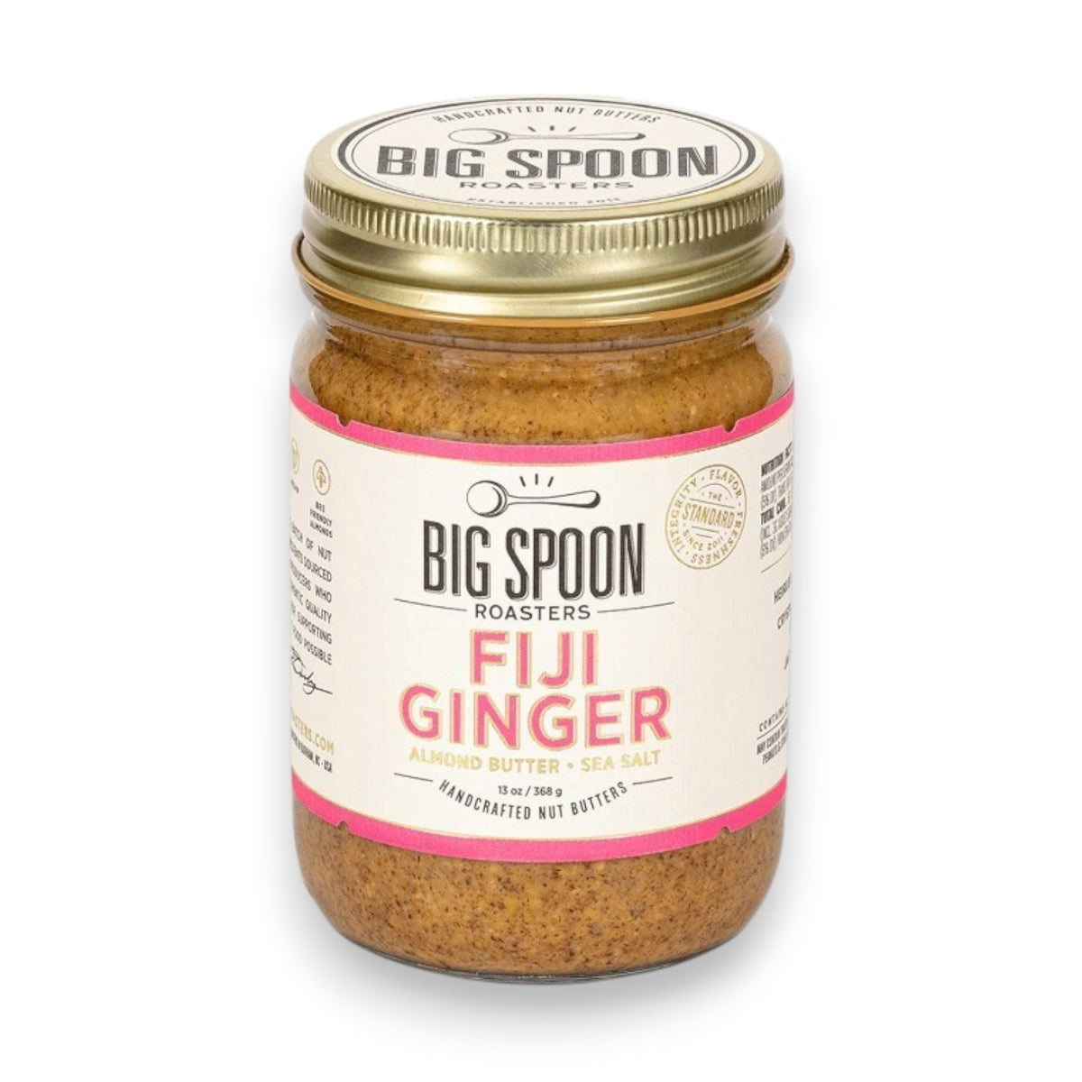 A jar of BIG SPOON ROASTERS - FIJI GINGER ALMOND, featuring a gold lid and a white label with pink accents that displays the brand name, flavor, and additional product information. The crystallized ginger-infused almond butter is visible through the clear jar.