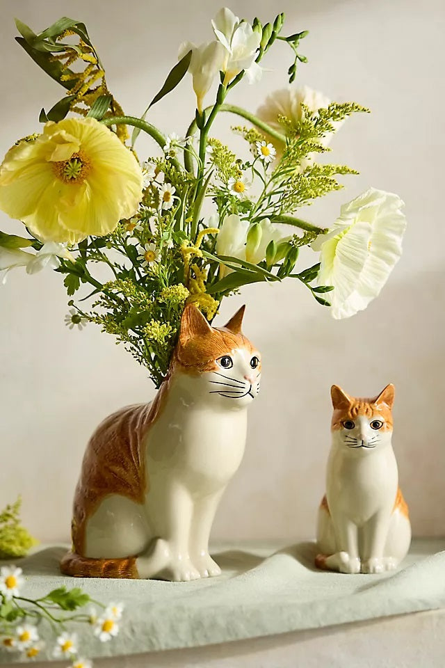 The QUAIL CERAMICS - SQUASH CAT FLOWER VASE by QUAIL DESIGNS is a hand-painted stoneware vase shaped like a sitting orange cat, with green foliage and yellow flowers appearing to grow from its back.