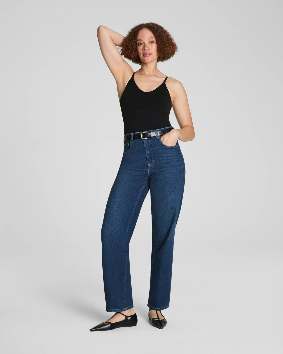 Wearing a black sleeveless top, SPANX 90&#39;S STRAIGHT LEG JEAN by SPANX, and black pointed-toe flats, a person stands against a plain light background.