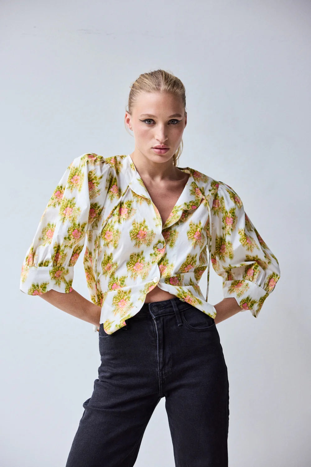 A woman with blonde hair in a ponytail wears the HOPE FOR FLOWERS Scalloped Shirt in Pink Warped Ikat by HOPE FOR FLOWERS and black high-waisted jeans, standing against a plain light background with her hands on her hips.