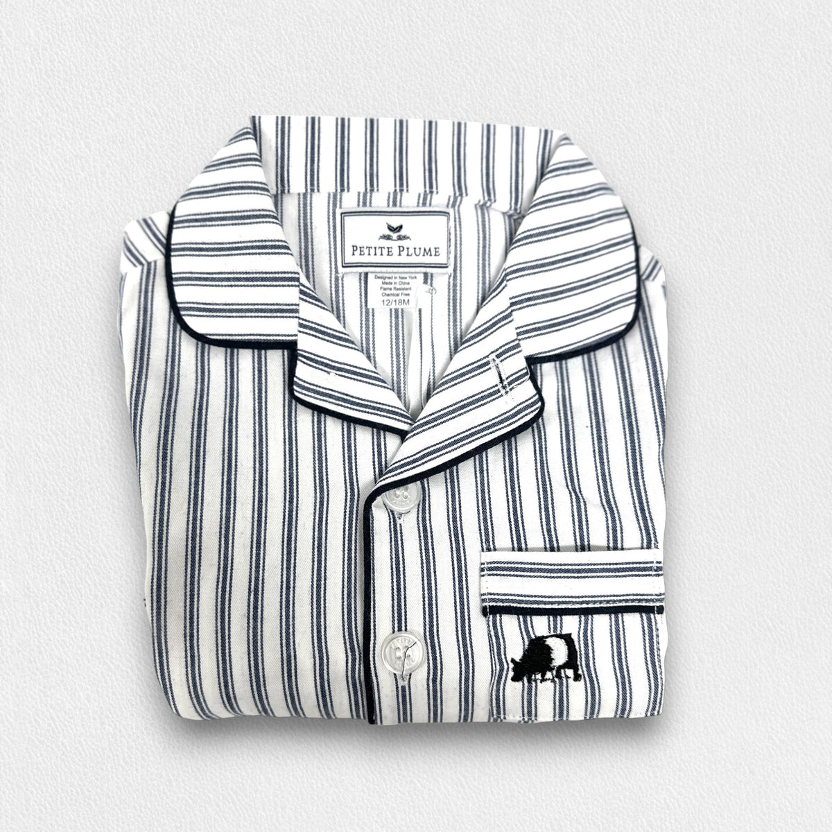 A young boy stands barefoot against a plain background, smiling and looking off to the side. He is wearing an elegant PETITE PLUME FEARRINGTON LIFESTYLE COLLECTION CHILDREN'S TWILL NAVY FRENCH TICKING PAJAMA SHORT SET and is holding a plush grey teddy bear by its arm.