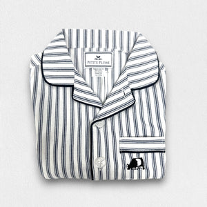 A young boy stands barefoot against a plain background, smiling and looking off to the side. He is wearing an elegant PETITE PLUME FEARRINGTON LIFESTYLE COLLECTION CHILDREN'S TWILL NAVY FRENCH TICKING PAJAMA SHORT SET and is holding a plush grey teddy bear by its arm.