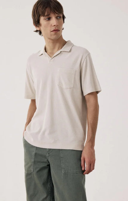 Wearing the HARTFORD Terry Polo Shirt in Stone with sage green cargo pants, a person stands with one hand in their pocket, glancing to the side against a plain background.