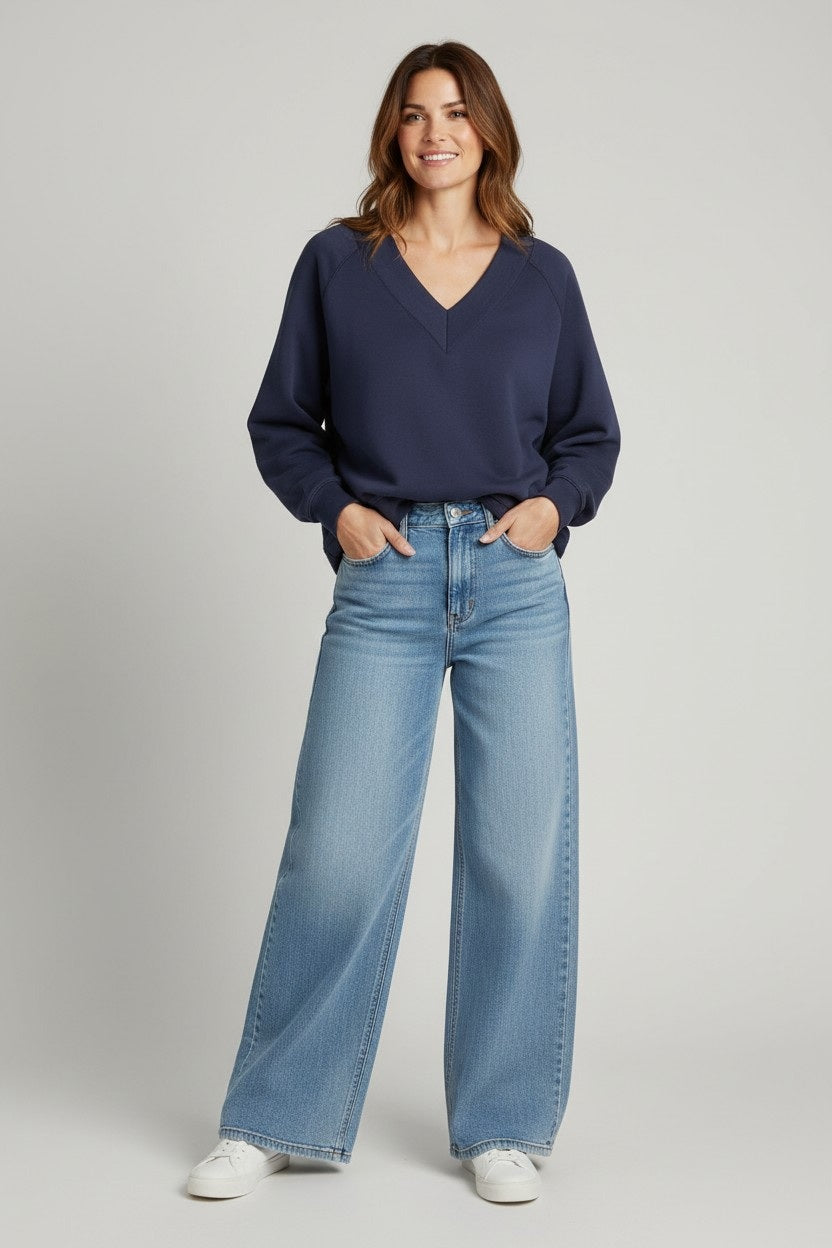 A woman stands smiling in a KMJ - TUCKER SWEATSHIRT by KMJ, featuring a relaxed fit and V-neckline in navy blue, paired with light blue high-waisted wide-leg jeans and white sneakers, hands in pockets against a light gray background.