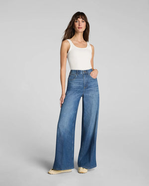 A person shown from mid-torso down against a plain gray background wears the SPANX SPANXSUPERSMOOTH Authentic Lightweight Super Wide Leg Jean in Oceanic paired with beige shoes.
