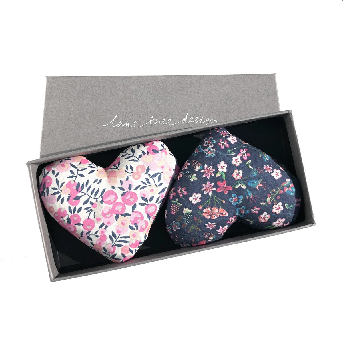 A grey rectangular box featuring the text &quot;LIME TREE COLLECTION LTD&quot; contains two heart-shaped fabric sachets. Branded as the HEARTS - LOVEY DOVEY SACHET, one sachet is adorned in Liberty Tana Lawn fabric with a pink, white, and blue floral pattern, while the other comes in dark blue with a small floral design. These delightful lavender drawer sachets will freshen up any space.