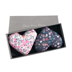 A grey rectangular box featuring the text "LIME TREE COLLECTION LTD" contains two heart-shaped fabric sachets. Branded as the HEARTS - LOVEY DOVEY SACHET, one sachet is adorned in Liberty Tana Lawn fabric with a pink, white, and blue floral pattern, while the other comes in dark blue with a small floral design. These delightful lavender drawer sachets will freshen up any space.