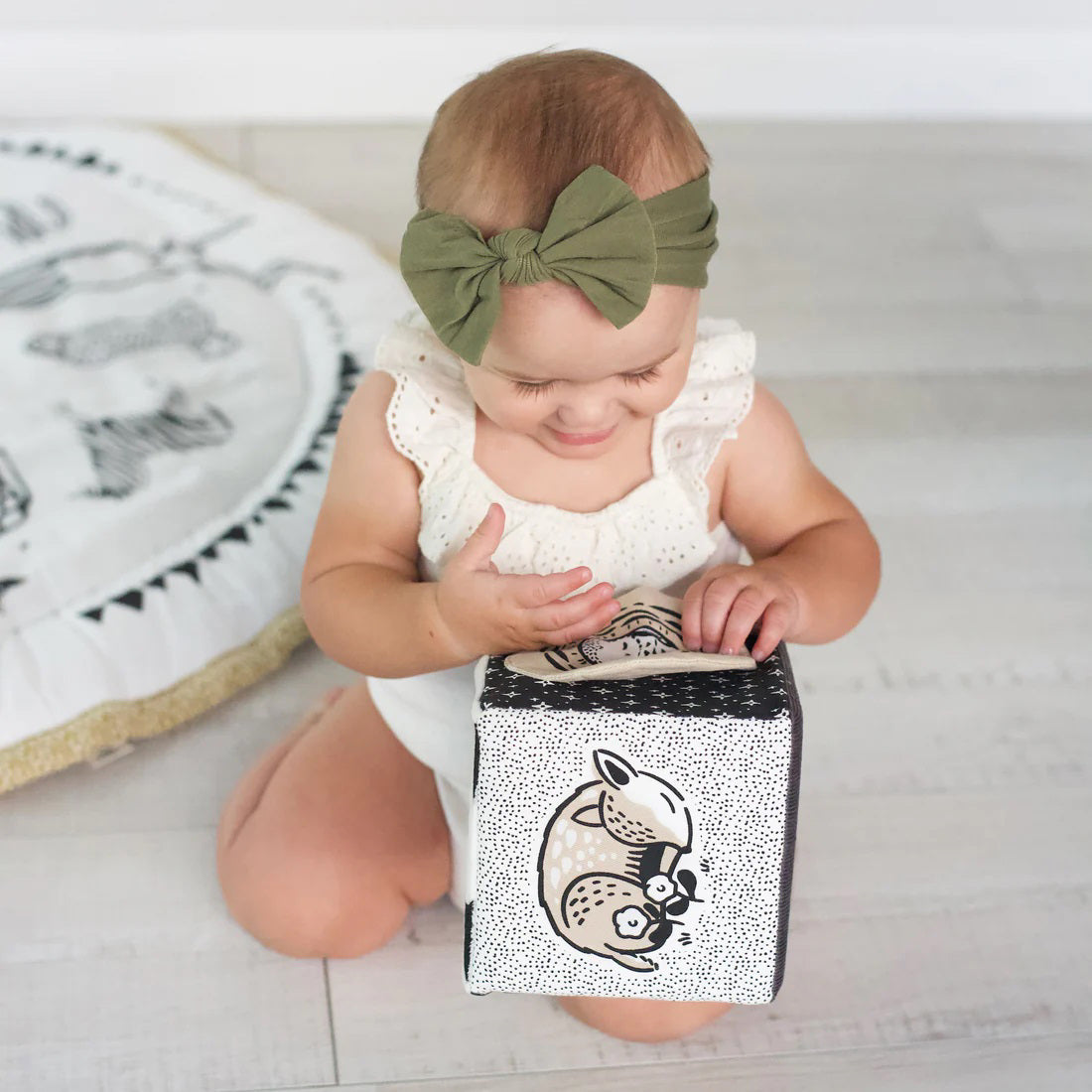 The WEE GALLERY - BABY ANIMALS SOFT BLOCK by WEE GALLERY is a fabric cube adorned with black and white designs that include adorable baby animals, such as a sleeping fox and its cub on one side. This sensory development toy features various textures, including polka dots and stripes, and includes two fabric loops on one edge to aid in tactile exploration.