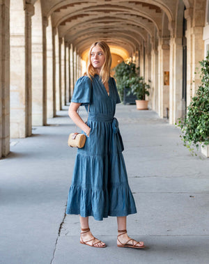 A woman with blonde hair, wearing the MERSEA Pariscope Maxi Dress in Chambray, stands by a stone wall. The blue denim maxi dress from MERSEA features short sleeves, a belted waist, and a subtle ruffle neckline for added elegance. She accessorizes with a gold necklace featuring a round pendant.