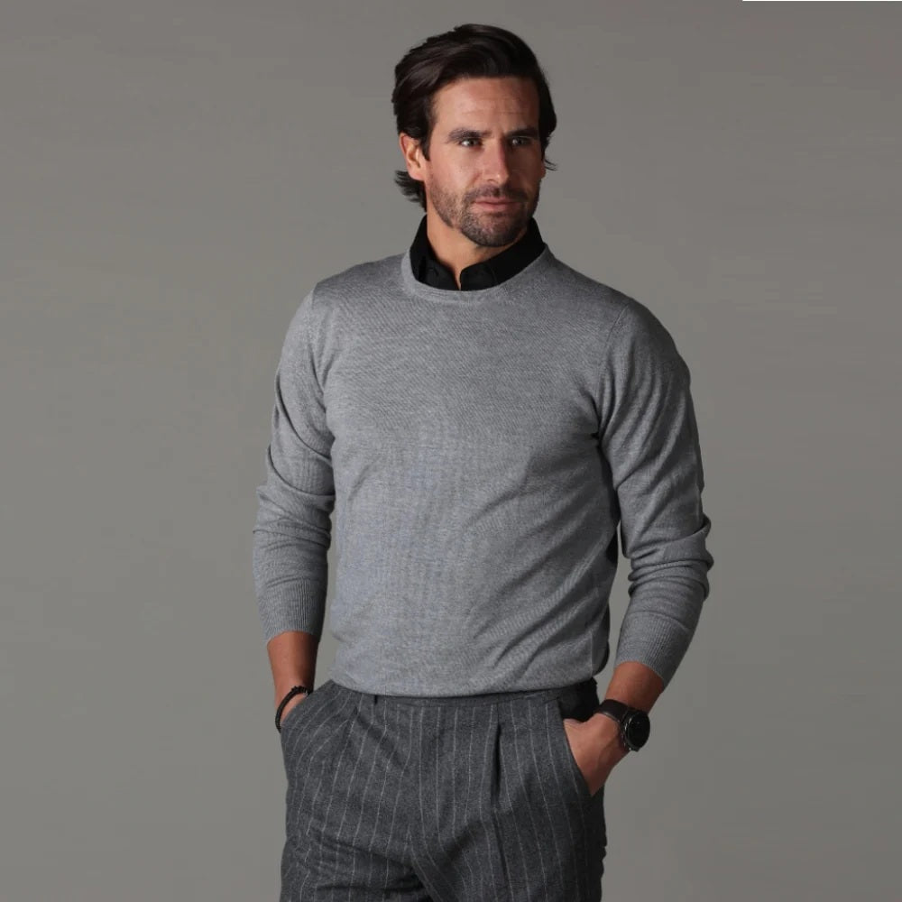 A man wearing the COLLARS AND CO - SEMI SPREAD COLLAR ORIGINAL POLO IN BLACK by COLLARS AND CO, paired with gray pinstripe pants, looks down while holding the hem of his shirt against a plain gray background.