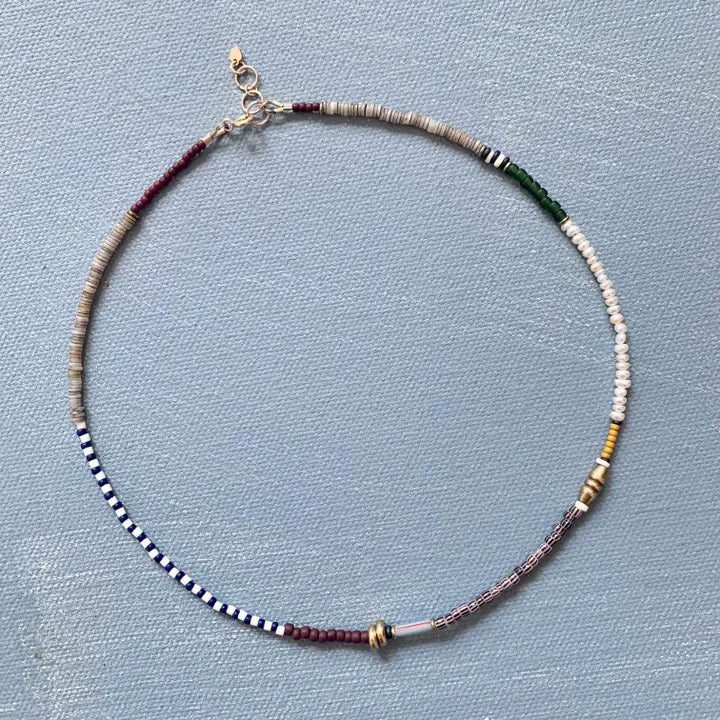 The ALICE RISE - PEARL MIXED MEDIA NECKLACE by ALICE RISE features small, multicolored vintage glass beads arranged in sections and finished with a 14k gold-filled clasp, displayed in a circle on a textured light blue surface.