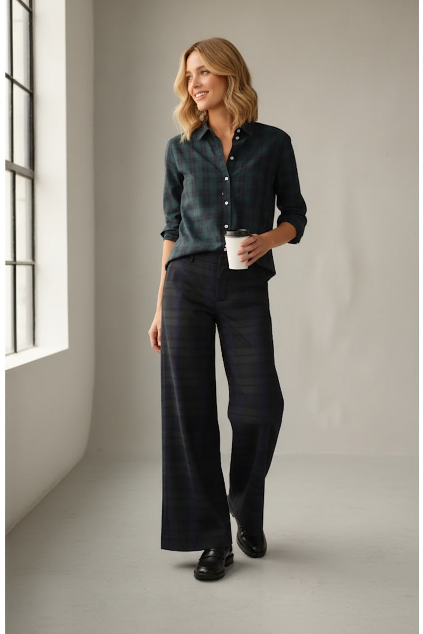 A woman stands indoors, smiling and holding a white coffee cup, wearing the FRANK & EILEEN West Village NYC Wide Leg Trouser in Black Watch Plaid as natural light streams in from large windows on her left.