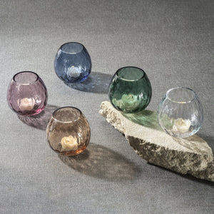 Five bronze PRISM Multipurpose Optic Vase Candleholders by ZODAX, each 4" x 3.75", hold white tealights and are arranged on a gray surface, with one displayed elevated on a rough stone slab.