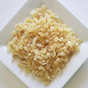 A close-up image of a square white plate containing small, beige-colored, crystal-like granules that appear to be THE CHAI BOX - CANDIED GINGER. The granules have a slightly translucent appearance.