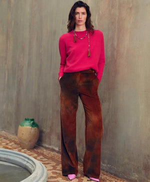 A person with long dark hair stands against a muted indoor wall, wearing a bright pink sweater, MOMONI - ISMAELTROUSER tie-dyed corduroy pants, and pink shoes. They have their hands in the patch pockets and accessorize with two long necklaces. An urn and a small water pool are placed beside them.