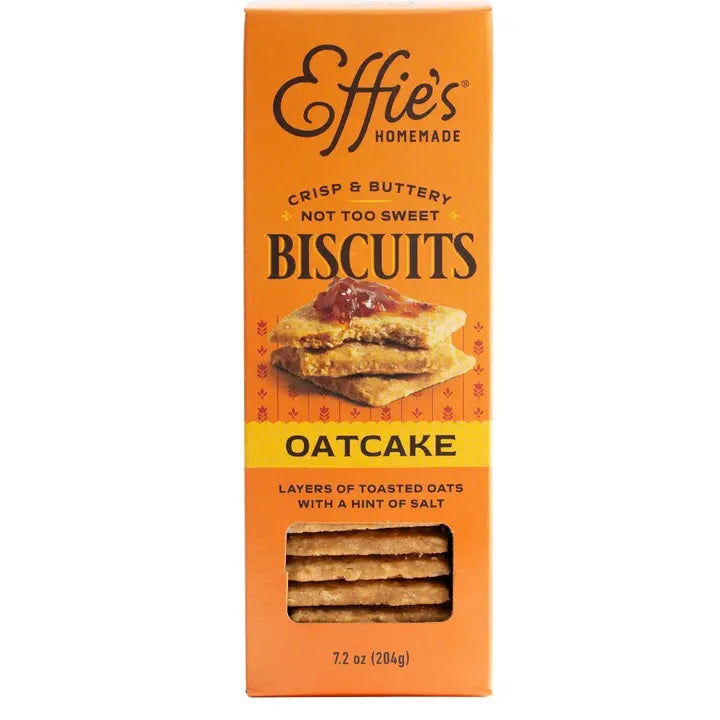 Two glass bowls filled with yogurt, blueberries, peach slices, and crumbled EFFIES - ORIGINAL OATCAKE BISCUIT by EFFIES HOMEMADE. A box of Effies Homemade Oatcake Biscuits, whole biscuits displaying their buttery layers, and golden spoons sit alongside. In the background, there’s a plate with peach slices.