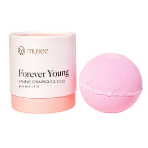 A pink bath balm made with natural ingredients sits next to its cylindrical container labeled “MUSEE - FOREVER YOUNG THERAPY BATH BALM” from MUSEE.
