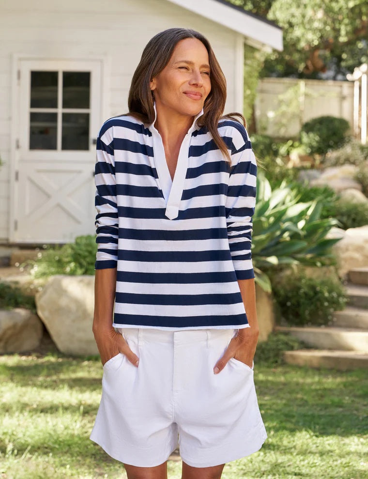 A woman stands outside, smiling, wearing the FRANK & EILEEN - Patrick Popover Henley in Vintage Jersey Rugby Nautical Stripe—a navy and white striped long-sleeve shirt crafted from 100% cotton—paired with white shorts. In the background are green plants and a white building with a window and door.