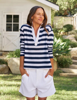 A woman stands outside, smiling, wearing the FRANK & EILEEN - Patrick Popover Henley in Vintage Jersey Rugby Nautical Stripe—a navy and white striped long-sleeve shirt crafted from 100% cotton—paired with white shorts. In the background are green plants and a white building with a window and door.