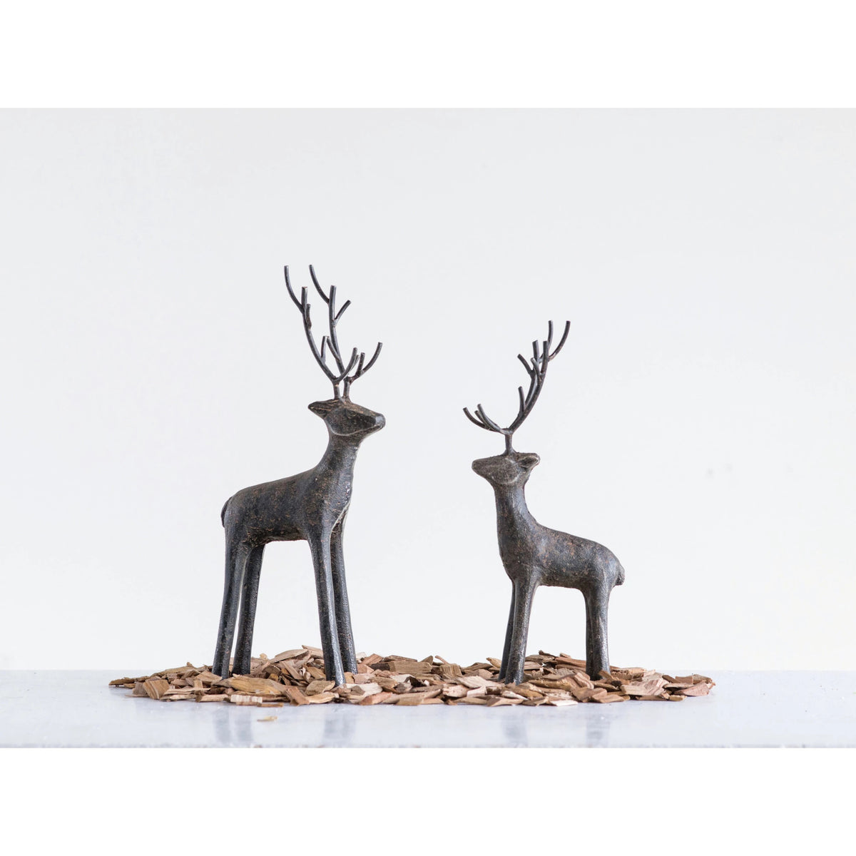 The CREATIVE COOP CAST IRON STANDING DEER features two minimalist black metal sculptures with tall antlers, displayed on wood chips against a white background, adding a striking modern touch to your holiday decor.