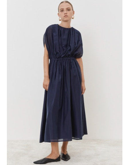 Wearing the ankle-length dark blue MEIMEIJ - OVERLAY DRESS, this person stands against a white backdrop. The dress features gathered sleeves and an elasticized waist, accessorized with black flats and large round earrings, while their hair is neatly pulled back.