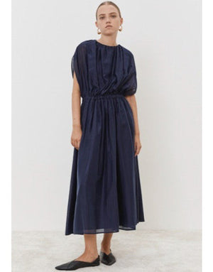 Wearing the ankle-length dark blue MEIMEIJ - OVERLAY DRESS, this person stands against a white backdrop. The dress features gathered sleeves and an elasticized waist, accessorized with black flats and large round earrings, while their hair is neatly pulled back.