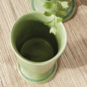 Two NAPA HOME AND GARDEN - KELCY FOOTED CACHEPOTS, finished with a green reactive glaze, rest on a light woven runner. One displays a leafy plant while the other is empty; a wooden chair and white slatted fence are seen in the background.
