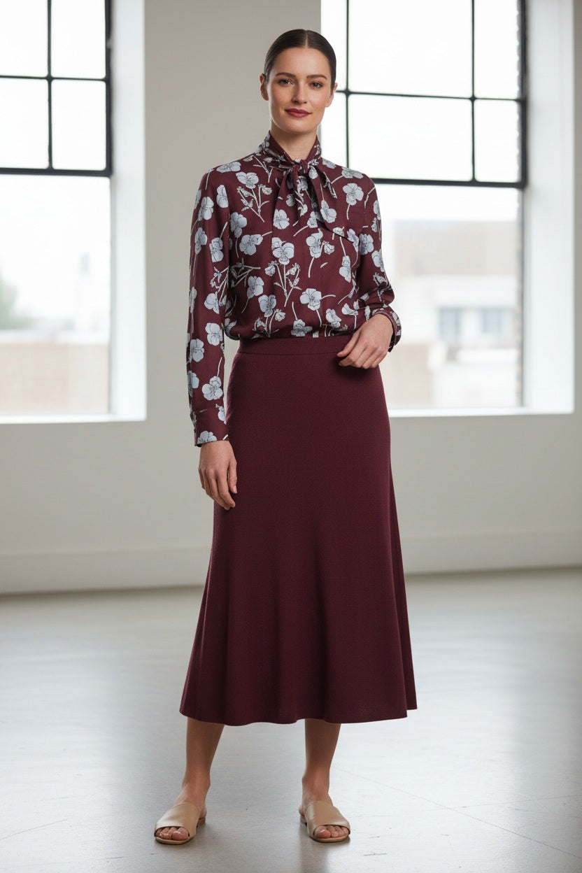 A woman stands indoors in front of large windows with blurred city views, smiling with her hair tied back. She is wearing a burgundy blouse with a white floral pattern, paired with the MIRTO 1956 - WOOL KNIT MIFI SKIRT IN BORDEAUX from MIRTO 1956, and beige sandals.