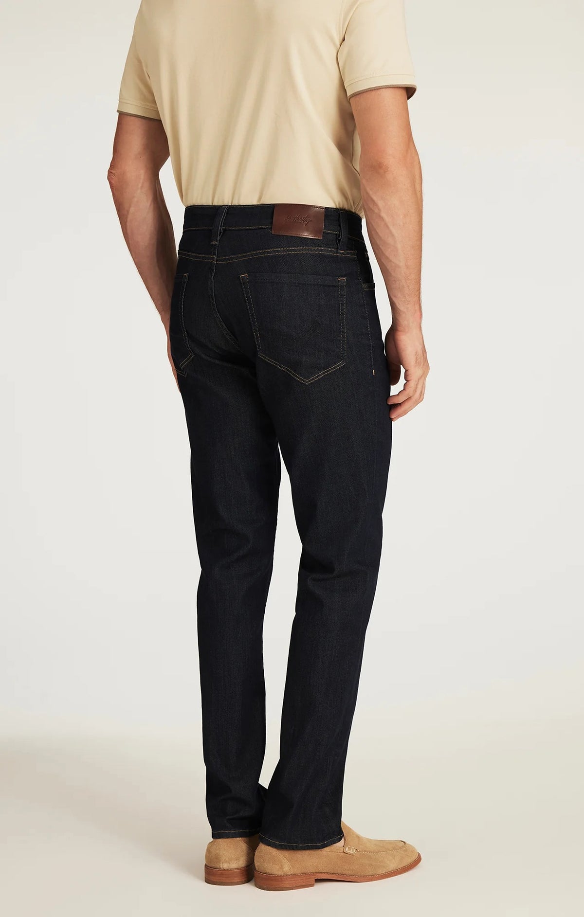 A person wearing 34 HERITAGE - CHARISMA RELAXED STRAIGHT JEANS IN RINSE VINTAGE, a dark blue, vintage-inspired denim by 34 HERITAGE, paired with a beige shirt and shown from the back. The jeans feature brown stitching and a brown leather patch on the waistband.