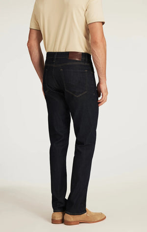 A person wearing 34 HERITAGE - CHARISMA RELAXED STRAIGHT JEANS IN RINSE VINTAGE, a dark blue, vintage-inspired denim by 34 HERITAGE, paired with a beige shirt and shown from the back. The jeans feature brown stitching and a brown leather patch on the waistband.