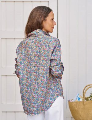 A woman with long brown hair smiles as she stands outside by a white wooden wall, wearing the FRANK & EILEEN EILEEN Relaxed Button Up in Italian Classic Linen Multicolor Flowers, paired with white pants and a straw tote bag nearby.