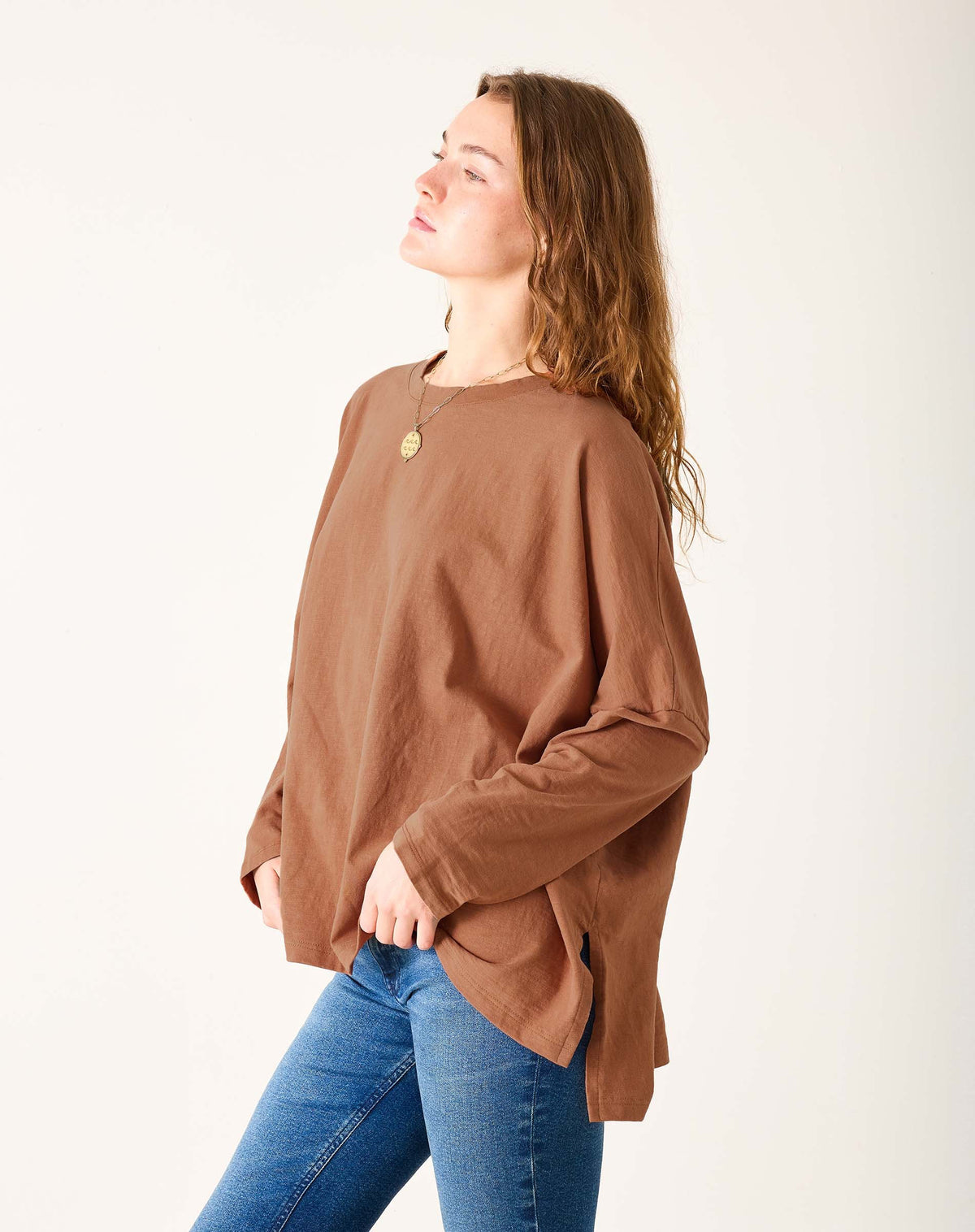 A woman with wavy brown hair, wearing the MERSEA Catalina Slub Tee in Suede and blue jeans, stands against a white wall, gazing to her right with a gentle smile.