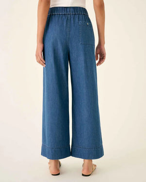 Wearing MERSEA - SAMMIE WIDE LEG PANT in light blue chambray with a pull-on waistband and front patch pockets, styled with a light top and cream sneakers, the person stands against a plain background.