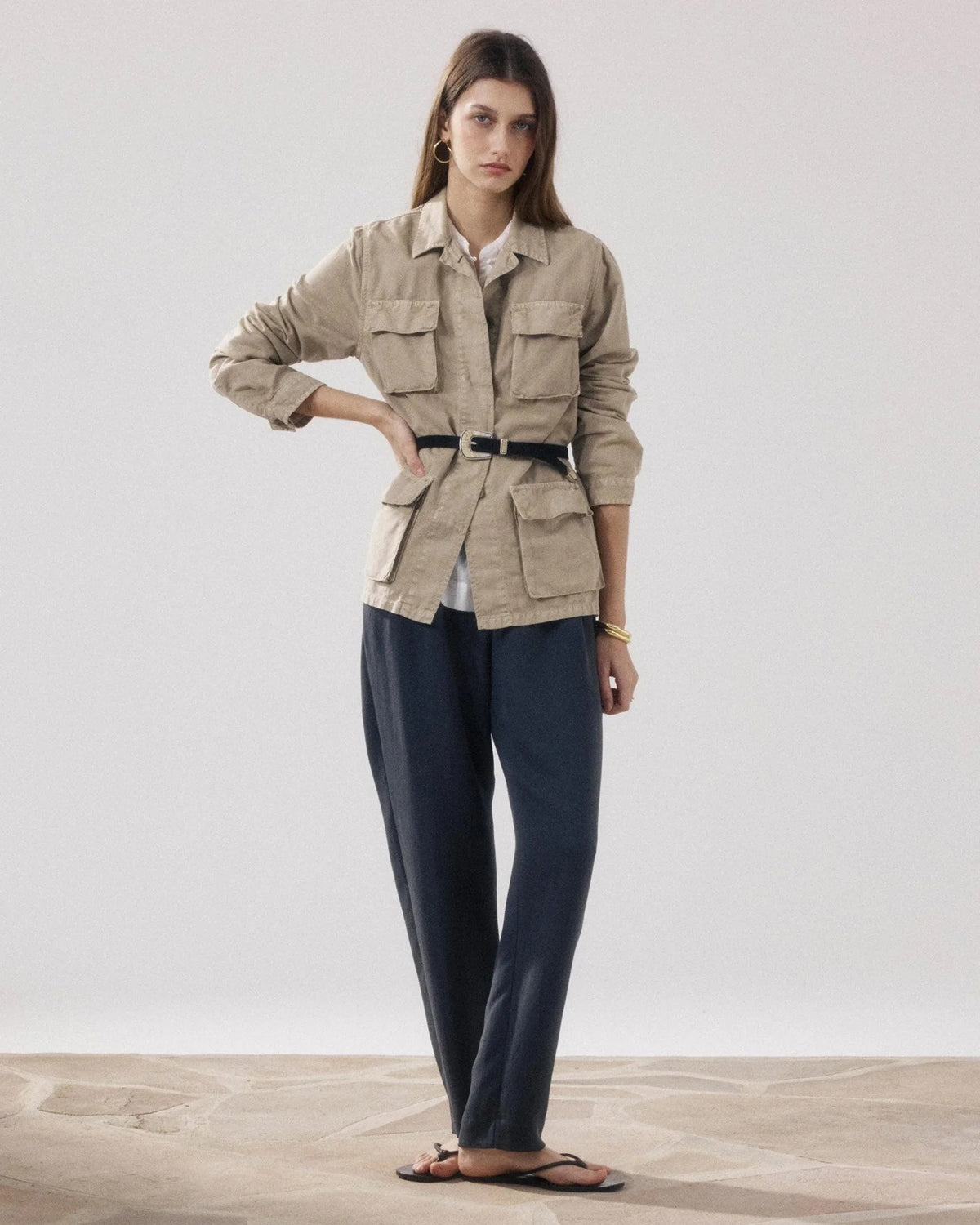 A woman stands indoors wearing the HARTFORD VEA Utility Jacket—a belted beige cotton-linen piece from HARTFORD—paired with dark loose pants, black sandals, and minimal jewelry. One arm is relaxed while the other is bent at the elbow. The background is plain and light.