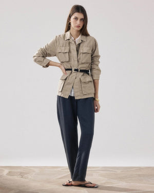 A woman stands indoors wearing the HARTFORD VEA Utility Jacket—a belted beige cotton-linen piece from HARTFORD—paired with dark loose pants, black sandals, and minimal jewelry. One arm is relaxed while the other is bent at the elbow. The background is plain and light.