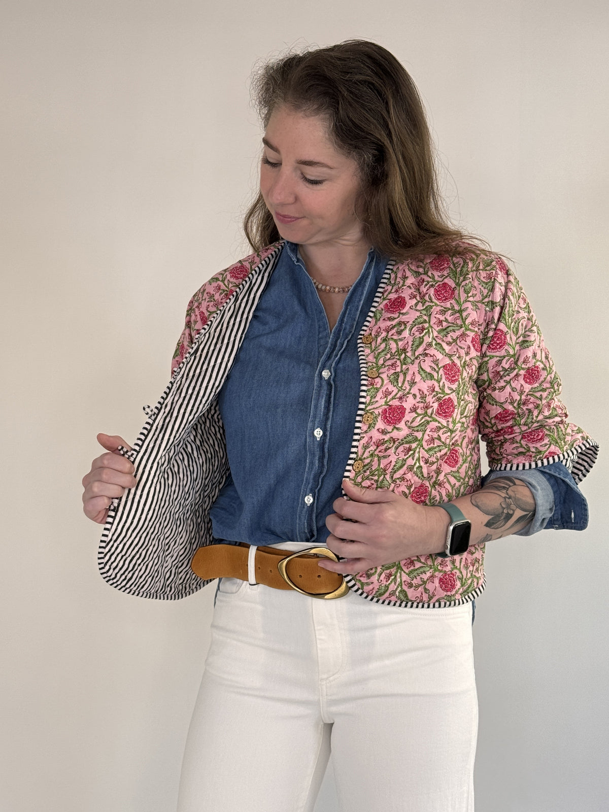 A woman with visible forearm tattoos smiles slightly against a plain background, wearing the FARYLINA ZOIE Reversible Quilted Jacket over a denim shirt, white pants, and a tan belt.