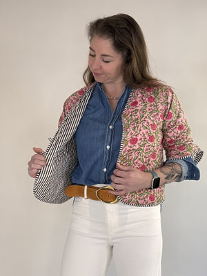 A woman with visible forearm tattoos smiles slightly against a plain background, wearing the FARYLINA ZOIE Reversible Quilted Jacket over a denim shirt, white pants, and a tan belt.