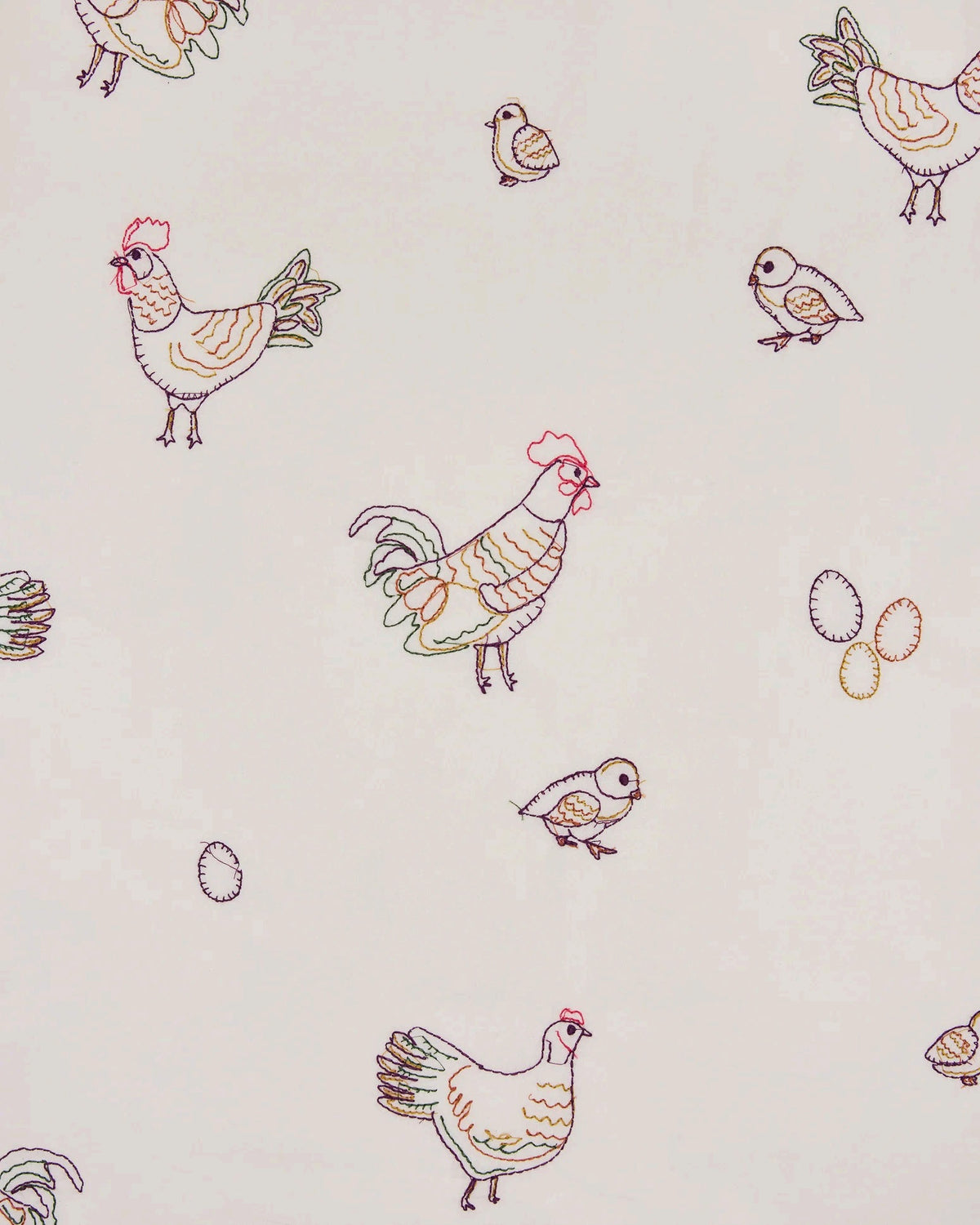 A woman dressed in the PRINTFRESH Embroidered Hens Long Pajama Set in Buff smiles as she stands by a wooden fence, holding a basket and watching chickens peck at the ground in a farmyard setting, with trees and a path visible behind her.