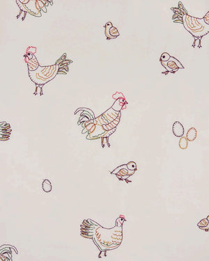 A woman dressed in the PRINTFRESH Embroidered Hens Long Pajama Set in Buff smiles as she stands by a wooden fence, holding a basket and watching chickens peck at the ground in a farmyard setting, with trees and a path visible behind her.