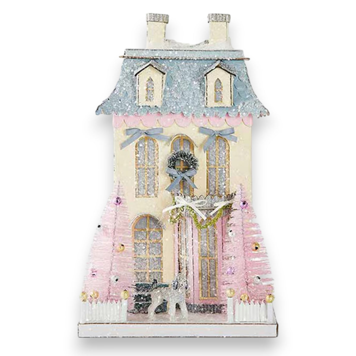 The CODY FOSTER WHITE CHATEAU HOLIDAY HOUSE features arched windows, a blue roof, pink accents, a wreath above the door, two pink ornamented trees, and a white picket fence—a charming collectible for design lovers.