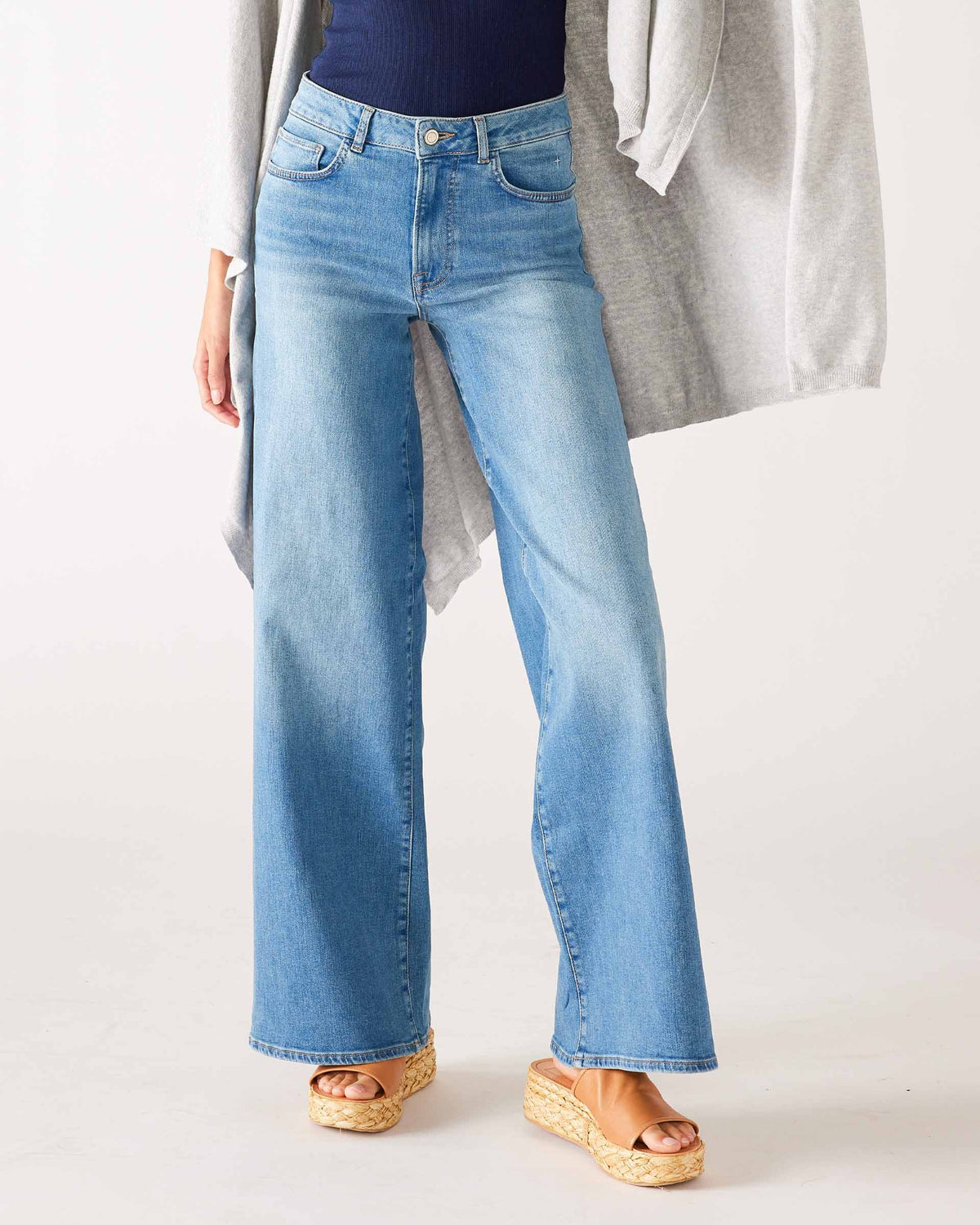 Someone stands against a neutral background wearing the MERSEA KATE WIDE LEG DENIM—light blue, high-waisted jeans made from recycled cotton—paired with a navy top, gray cardigan, and tan platform sandals.
