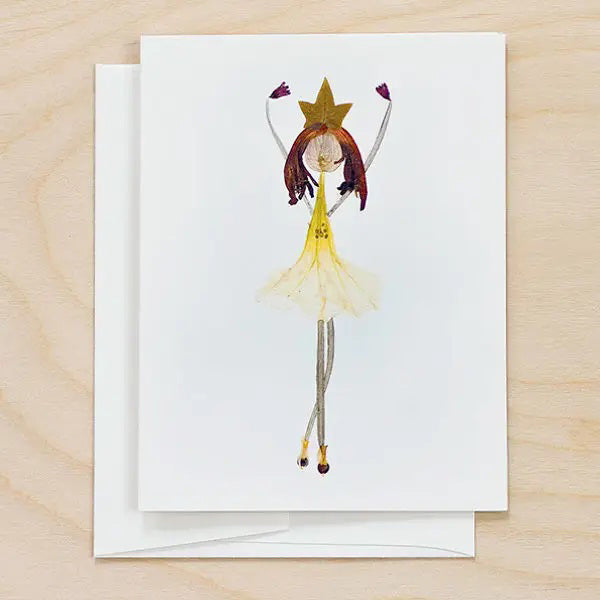 The PETAL PEOPLE PRESS - BALLERINA CARD by PETAL PEOPLE PRESS features a girl in a yellow dress and star headpiece, perfect as a thank you note card. Shown on a white surface with green leaves, scissors, and paperclips.