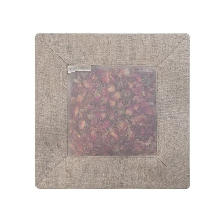 A SQUARE ROSE SACHET IN NATURAL LINEN by E WIGHTMAN &amp; CO features a beige linen square filled with dried rose petals visible through a translucent center, displayed on a plain white background. The delicate pink and red petals create an elegant floral display through the fabric window, providing both beauty and clothing protection.