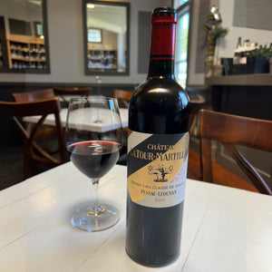A bottle of Château Latour-Martillac Rouge 2019 by Monsieur Touton features a yellow label with text and a building image. The neck is wrapped in red foil, evoking lush black fruit flavors against a plain white backdrop.