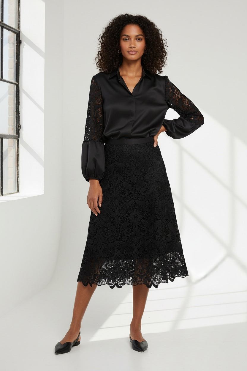 A woman stands against a plain background, wearing a black satin blouse with lace sleeves, the HINSON WU - GLORIA LACE SKIRT by HINSON WU, and chic pointed-toe black shoes, with one hand raised near her ear.