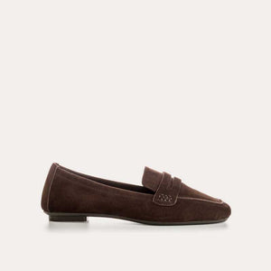 A person stands on a light floor wearing rolled-up blue jeans and REQINS HEMA suede loafers in Marron, showcasing the artisanal craftsmanship of REQINS.