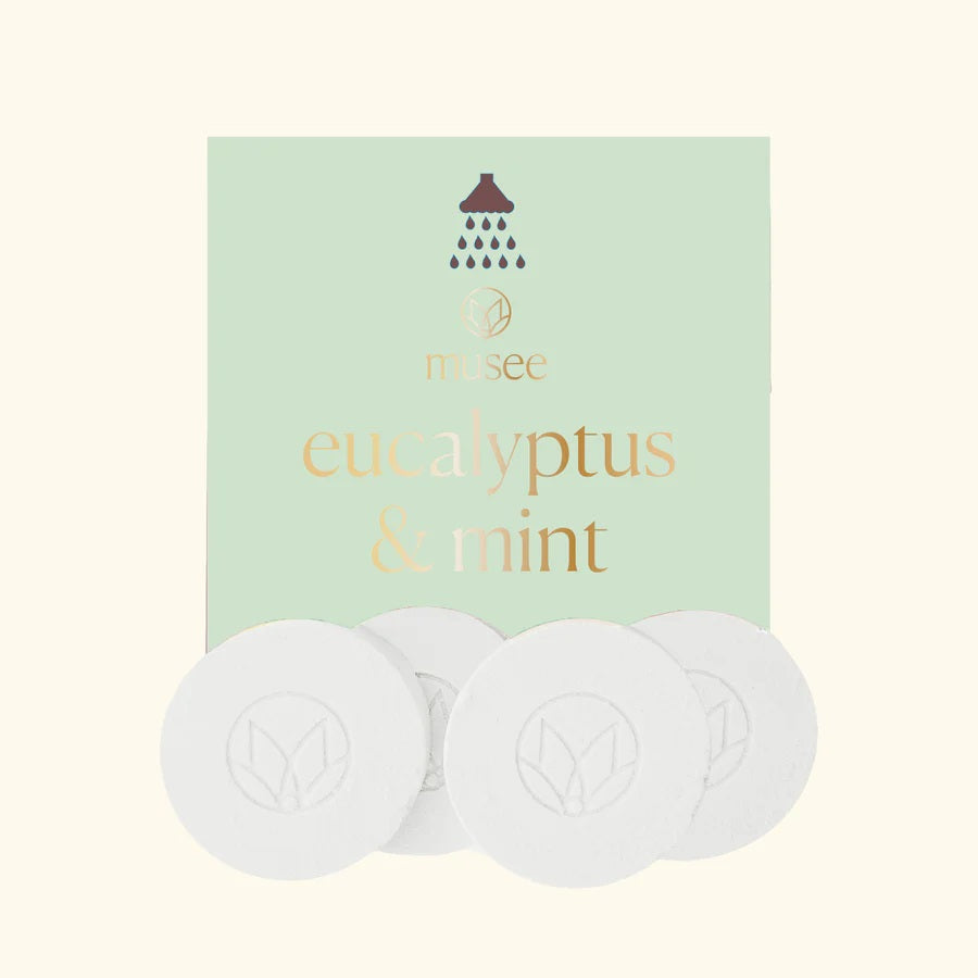 A pastel green packet labeled “MUSEE - EUCALYPTUS & MINT SHOWER STEAMERS” from MUSEE, featuring a shower icon, is displayed behind four round white shower steamers embossed with a leaf design—offering a spa-like experience with these natural bath products.