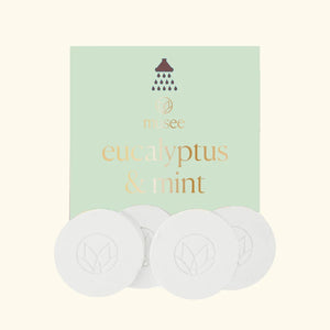 A pastel green packet labeled “MUSEE - EUCALYPTUS & MINT SHOWER STEAMERS” from MUSEE, featuring a shower icon, is displayed behind four round white shower steamers embossed with a leaf design—offering a spa-like experience with these natural bath products.