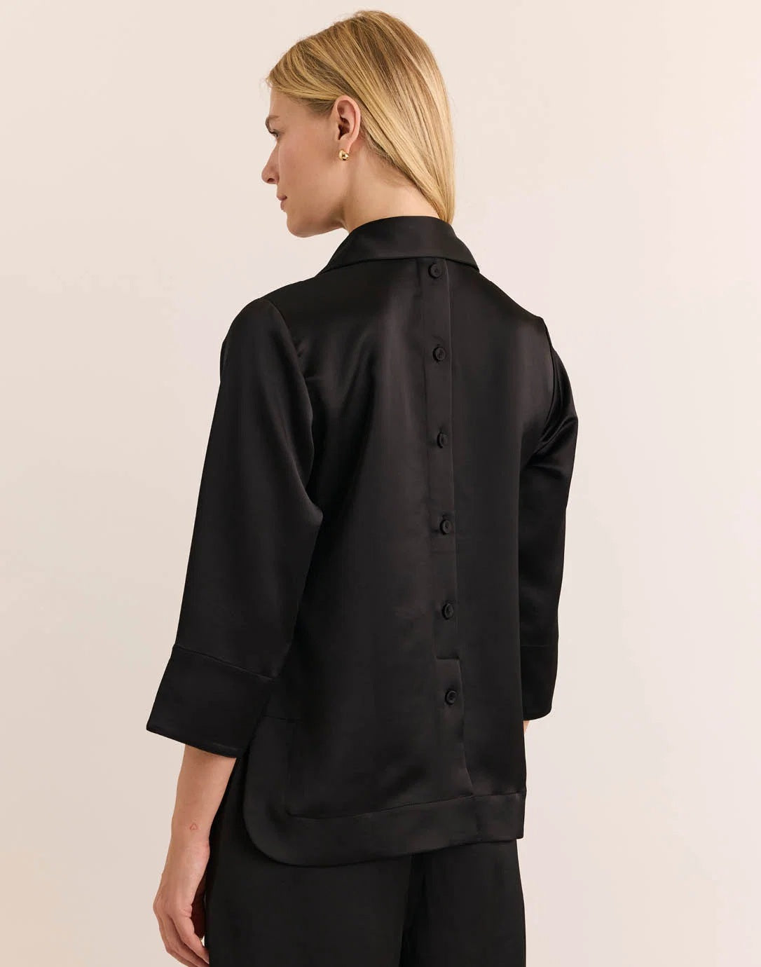 A woman with blonde hair models the HINSON WU Aileen 3/4 Sleeve Liquid Satin Shirt in black, featuring a collar and button details, against a plain light background.