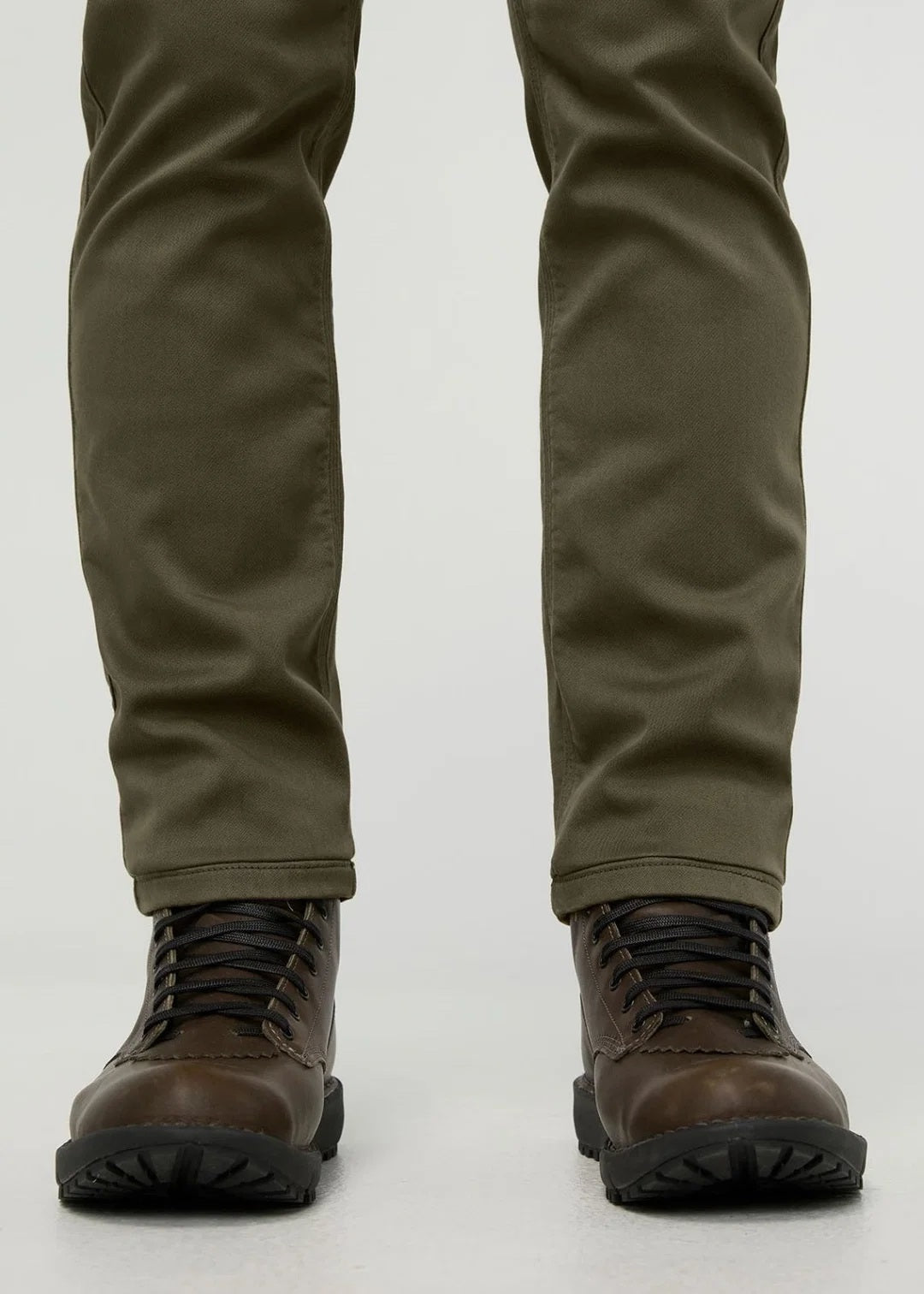 A person stands facing away from the camera against a plain background, wearing DUER - NO SWEAT ATHLETIC TAPER PANTS IN KELP paired with brown shoes.